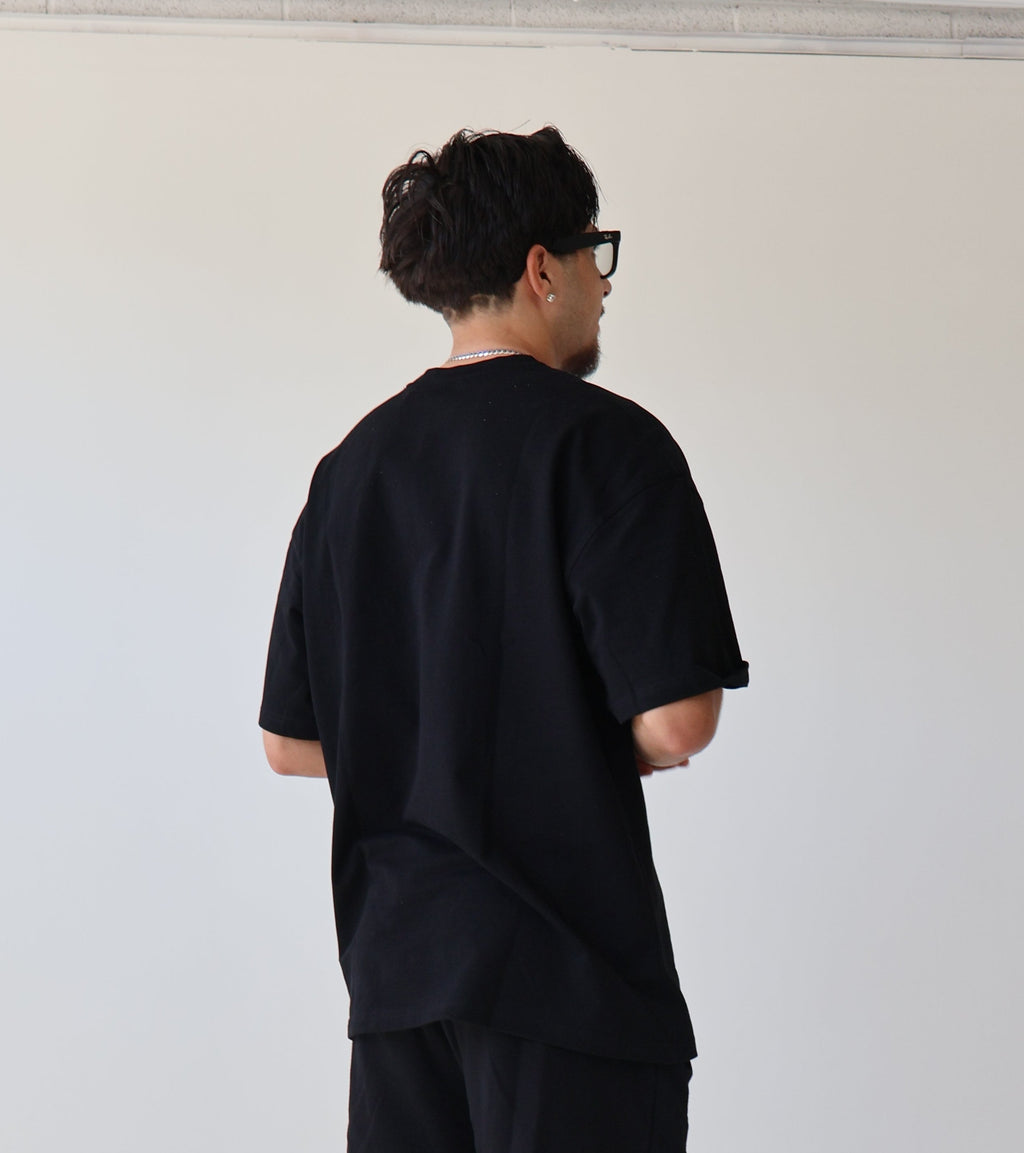 A man stands with his back to the camera wearing a loose black t-shirt, black shorts, black socks, and chunky black and white sneakers in a minimalist studio setting.