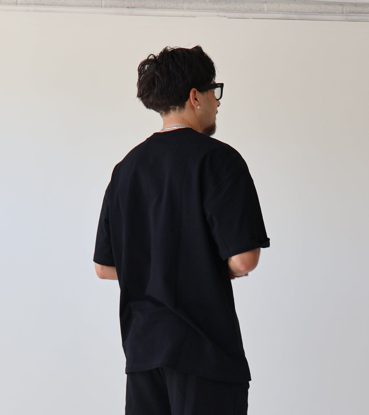 A man stands with his back to the camera wearing a loose black t-shirt, black shorts, black socks, and chunky black and white sneakers in a minimalist studio setting.