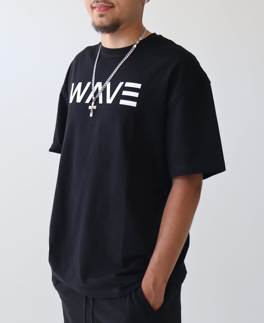 A man wearing a black oversized t-shirt with 'WAVE' printed in white across the chest, paired with black pants, a silver chain necklace, and glasses, standing against a plain light gray background.