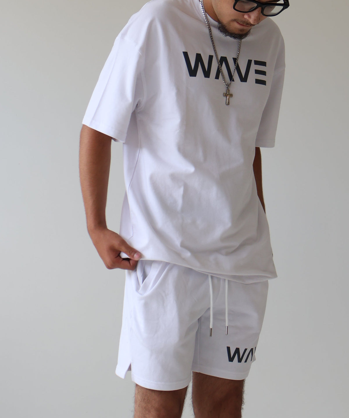 A man wearing a white oversized t-shirt with 'WAVE' printed on the chest and matching white shorts, standing in a minimalist white studio.