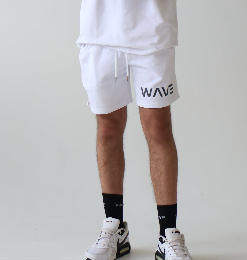 A man wearing a white t-shirt and matching white shorts with black 'WAVE' text logos, standing in a bright studio with a white wall background.