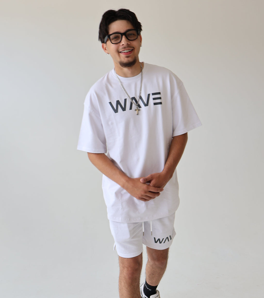 A man wearing a white oversized t-shirt with 'WAVE' printed on the chest, matching white shorts, white sneakers, and black socks, standing in a bright white studio.