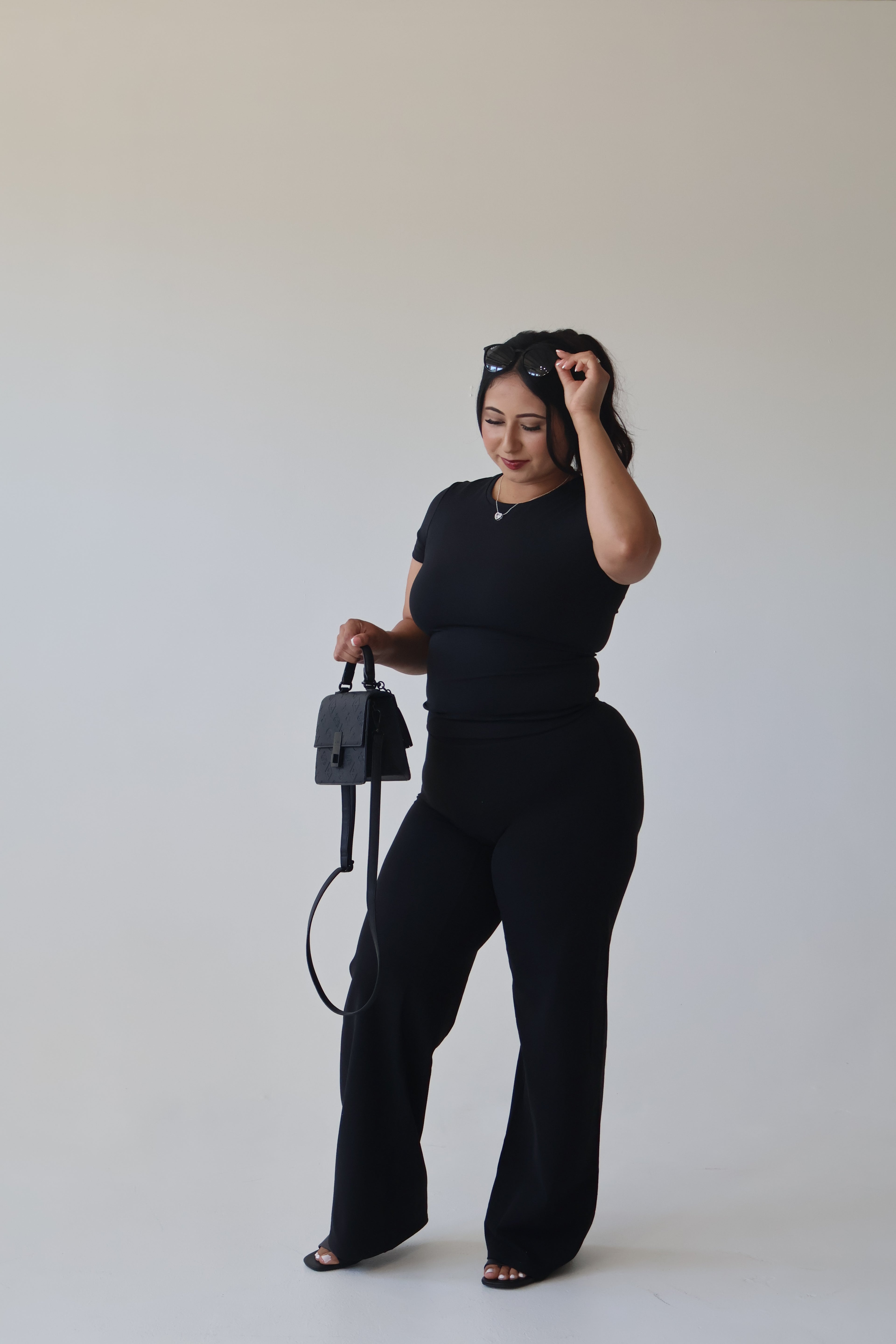 Woman in black outfit holding a black bag against a plain background