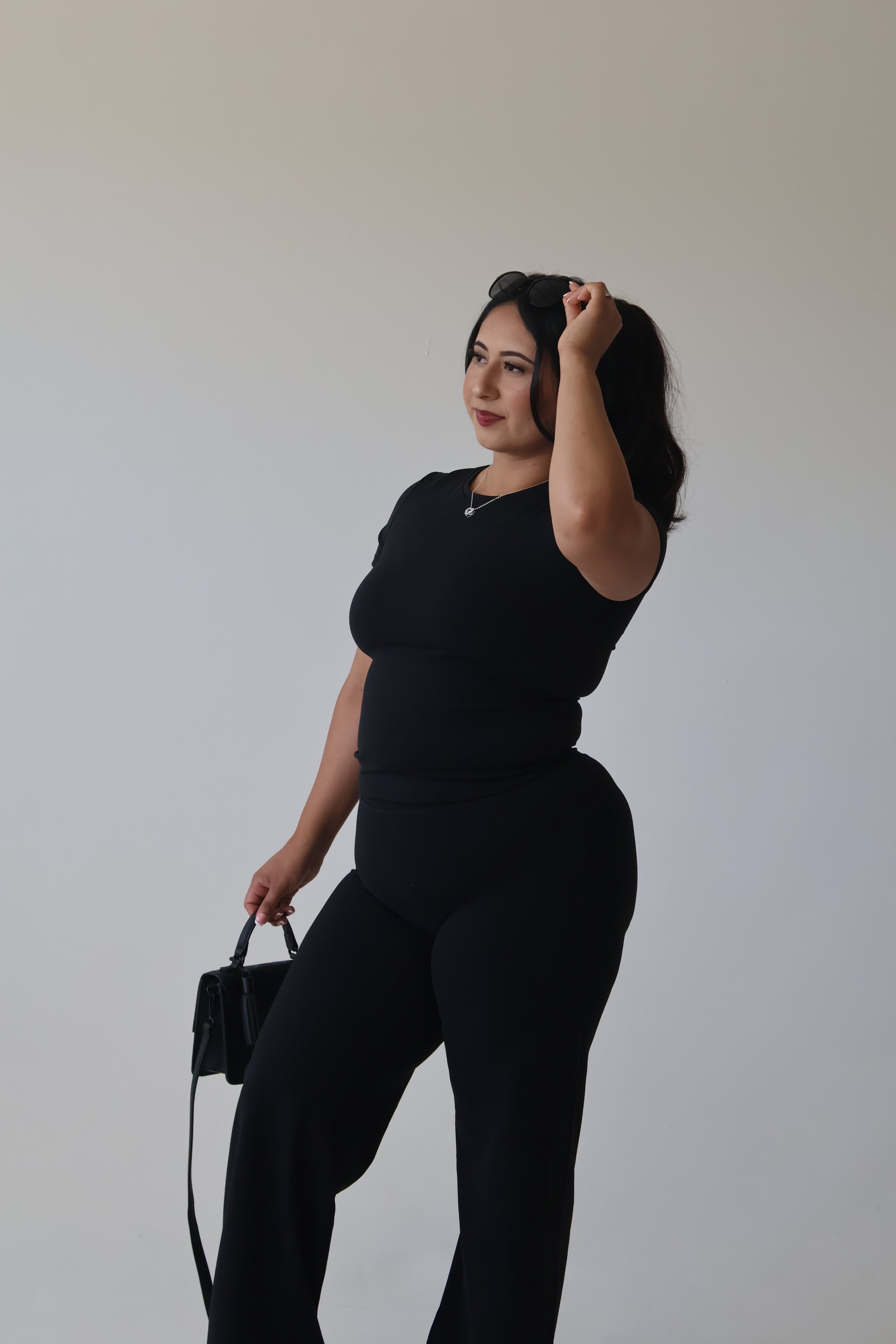 Woman in a black outfit holding a black handbag against a plain background