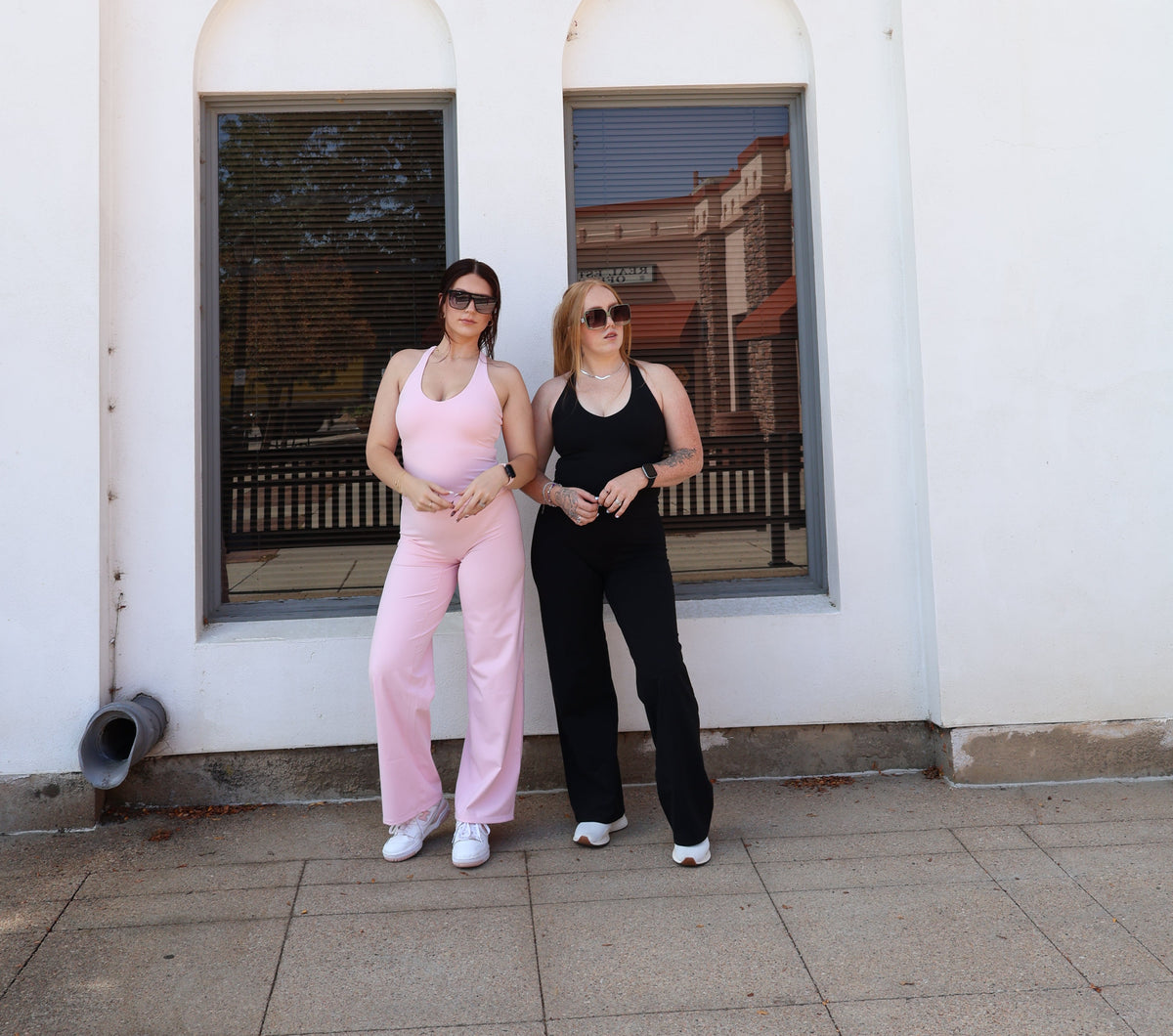 Two women posing in front of a white building with arched windows. The woman on the left wears a light pink sleeveless jumpsuit and white sneakers, while the woman on the right wears a black sleeveless top and black pants.